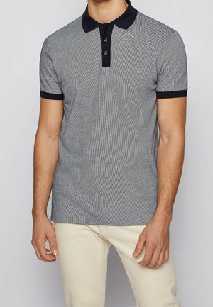 Mężczyzna w szarej koszulce polo z krótkim rękawem, czarnym kołnierzykiem i mankietami, w jasnobeżowych spodniach, stojący na jednolitym tle.