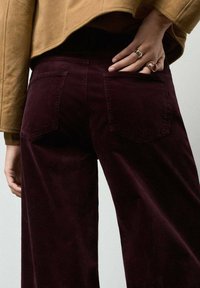 Pantalones de pana burdeos con piernas anchas, que cuentan con dos bolsillos traseros y una superficie texturizada con estrías, combinados con una chaqueta marrón corta.