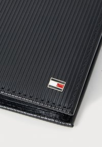 Carteira texturizada preta com sulcos verticais, apresentando um pequeno logotipo metálico em vermelho, branco e azul. Forma retangular compacta.