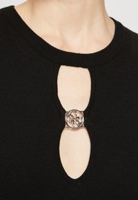 Top negro de punto con escote en forma de ojo de cerradura, acentuado por un broche circular en tono plateado en el centro. Textura suave con un ribete acanalado.