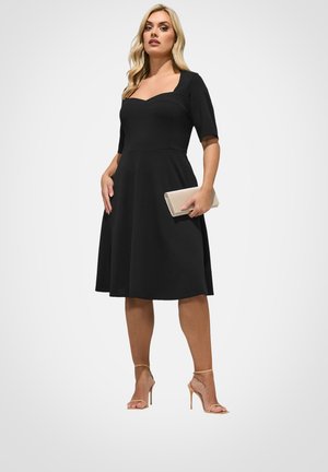 Woman wearing a knee-length black dress with short sleeves, holding a beige clutch, and wearing beige high-heeled sandals.