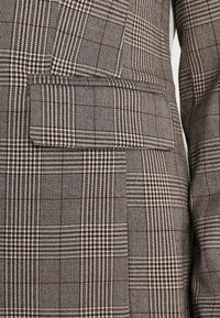Hnědo-beige kostkovaný blejzr, který má texturovanou tkaninu, jedno prsní kapsu a kombinaci vzorů houndstooth a kostkovaného.