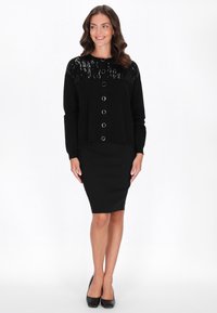 Cardigan nero con parte superiore testurizzata, scollo rotondo e sette bottoni in tono argento. Abbinato a una gonna a tubino nera aderente e tacchi.