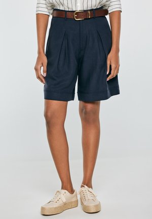 Kobieta ubrana w granatowe plisowane szorty z wysokim stanem i brązowym paskiem, białą koszulę w paski oraz kremowe trampki na platformie, stojąca na jednolitym tle.