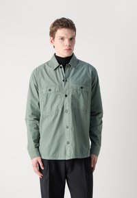Camisa de manga larga verde claro con frontón abotonado, dos bolsillos en el pecho y un corte relajado. Confeccionada con una tela suave y ligera.