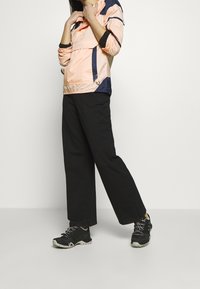 Ljus persikofärgad och marinblå vindjacka med dragkedja framtill och elastisk nederkant, kombinerad med svarta vida byxor och svarta sportiga skor.