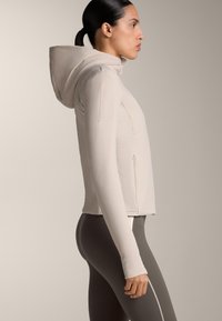 Ženska s temnimi lasmi v čop, oblečena v svetlo bež jakno s kapuco in temnosive pajkice z vertikalno svetlo črto, stoji postrani.