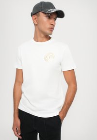 Hvit bomull t-skjorte med korte ermer og rund hals, med et gulllogo detalj på brystet. Sammen med mørke bukser og en grå caps.