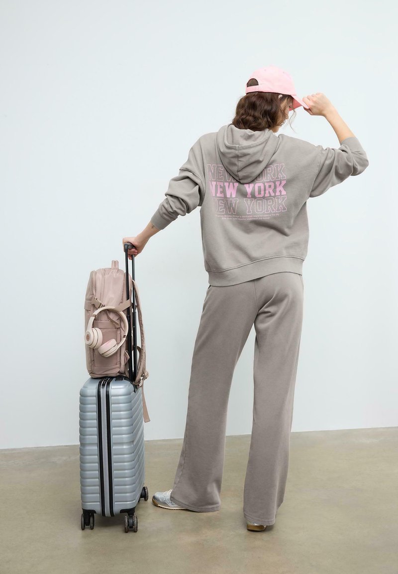 Person i gråt træningssæt og lyserød kasket, der holder en sølvfarvet kuffert med hjul, hvor en lyserød rygsæk og hovedtelefoner er fastgjort, stående mod en hvid væg.