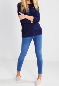 Blondin seisoo kädet ristissä, päällään tummansininen pitkähihaisten paita, vaaleansiniset kapeat farkut ja vaaleanpunaiset liukukengät.