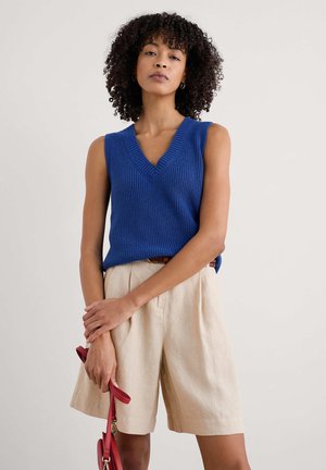 Top senza maniche blu in maglia con scollatura a V, abbinato a pantaloni corti in lino beige. La modella tiene una borsa rossa, mostrando un look casual.