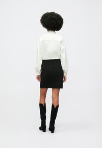 Pessoa com cabelo afro vestindo uma blusa de mangas compridas branca, saia preta e botas altas pretas, de pé contra um fundo simples.