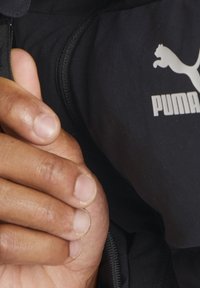 Veste Puma noire avec une poche zippée. Gros plan sur une main reposant sur le tissu, mettant en valeur sa texture lisse et son ajustement.