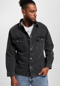 Svart denimskjorte med knappelukking, to brystlommer og lange ermer. Har krage og kontrasterende sømadetaljer.