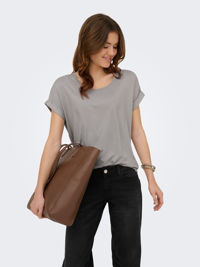 Jeune femme en t-shirt gris et pantalon noir tenant un grand sac en cuir marron, souriante et regardant vers le bas.
