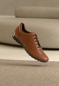 Καφέ δερμάτινο παπούτσι με λεία υφή, διαθέτει στρογγυλεμένη μύτη, σχέδιο με κορδόνια και μαύρη λαστιχένια σόλα. Διακριτική λεπτομέρεια ραφής ορατή.