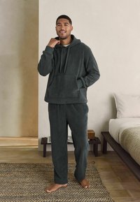 Completo loungewear verde scuro realizzato in tessuto testurizzato, composto da una felpa con cappuccio e tasca frontale e pantaloni larghi.