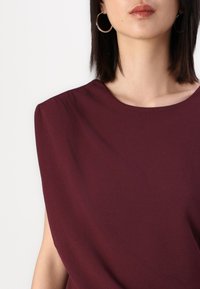 Burgundská blúzka s hladkou textúrou, okrúhlym výstrihom a skladmi na ramenách. Skombinovaná s veľkými kruhovými náušnicami, čiastočne viditeľný kľúčový kost.