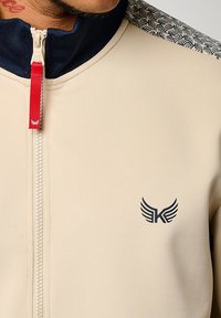 Veste zippée beige avec un col haut, des accents bleu marine, une tirette rouge et un logo ailé noir sur la poitrine. Motif texturé sur les épaules.