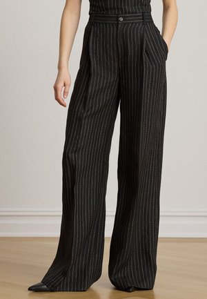 Personne portant un pantalon noir taille haute à jambes larges à rayures fines, les mains dans les poches, debout sur un sol en bois contre un mur uni.