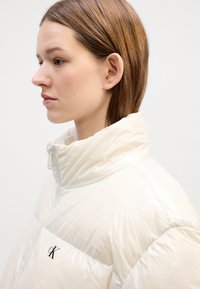 Jakna od punjenog materijala u sjajnoj, off-white boji s visokim ovratnikom. Ima zatvarač i crni logo na prsima, s kviltanim dizajnom.