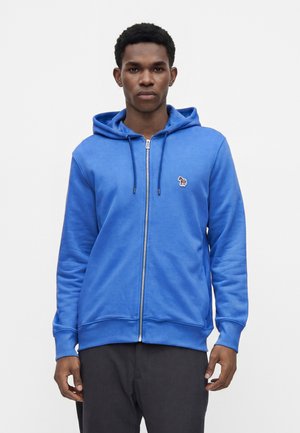 Jeune homme portant un sweat à capuche bleu zippé avec un petit logo zèbre sur la poitrine et un pantalon noir sur un fond uni.