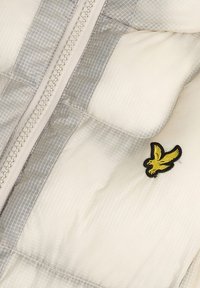 Gros plan d’une veste matelassée blanche avec des accents gris, une fermeture éclair et un petit emblème d’oiseau jaune brodé sur le tissu.