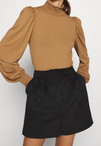 Braunes Langarmoberteil mit rüschigem Hochauschnitt und Puffärmeln, kombiniert mit hochgeschnittenen schwarzen Shorts aus einem weichen, strukturierten Stoff.