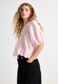 Světle růžová, poloprůsvitná blůza s kulatým výstřihem, nabíranými rukávy a volánovým lemem. Spárována s černými džíny s vysokým pasem.