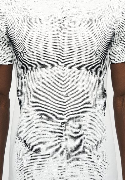 Κοντινό πλάνο ενός κορμού που φοράει λευκό μπλουζάκι με μαύρο βουλωτό και γραμμωτό σχέδιο που μοιάζει με αφηρημένο ανατομικό στήθος και κοιλιά.