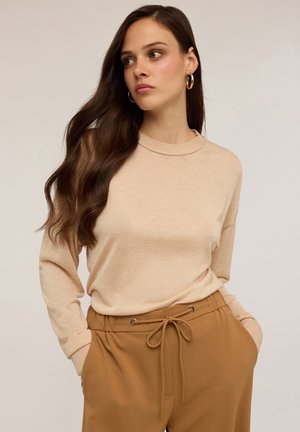 Maglione beige a maniche lunghe con scollatura rotonda, texture morbida e vestibilità comoda, abbinato a pantaloni beige con coulisse e occhielli metallici.
