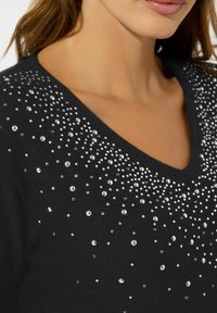 Schwarzer V-Ausschnitt-Pullover mit verstreuten klaren Strasssteinen in unterschiedlichen Größen über der Brust, die einen strukturierten, schimmernden Effekt erzeugen.