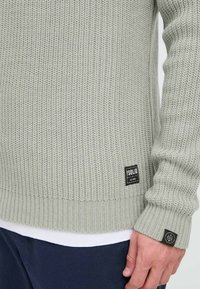Lys grå strikket genser med ribbet tekstur, har en svart merkeetikett og en ekstra logo-accent på mansjetten. Casual passform.