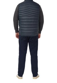 Dunkelblaue Steppweste mit einem Zickzack-Quiltmuster, getragen über einem grauen Langarmshirt, kombiniert mit dunklen Hosen und hellen Sneakern.