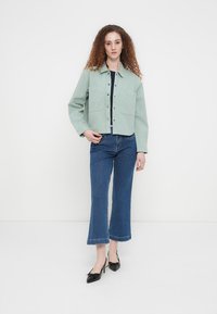 Veste courte vert clair avec poches avant, boutons argentés, associée à un jean bleu à jambes larges et des ballerines noires pointues.