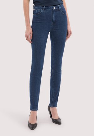 Schmal geschnittene dunkelblaue Jeans aus Denim mit Vorderentaschen und Knopfverschluss, getragen mit schwarzen High Heels vor hellem Hintergrund.