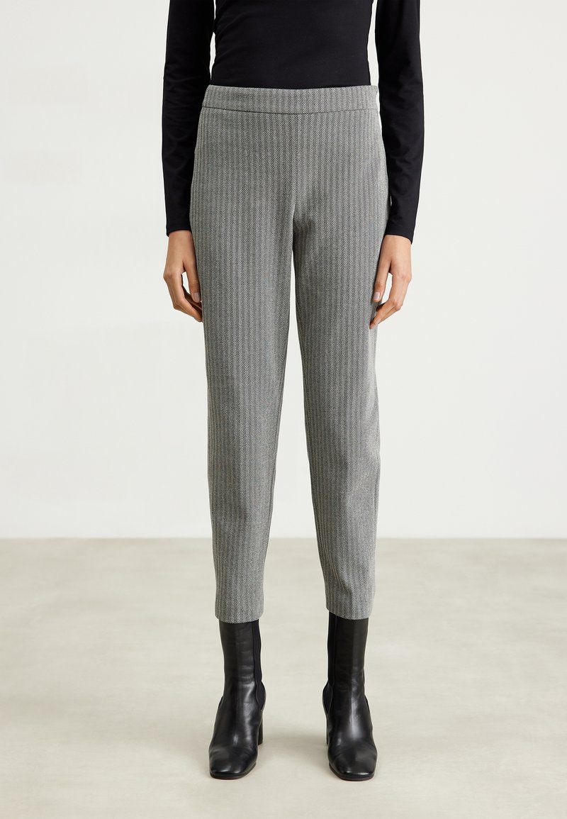 Femme portant un pantalon gris slim longueur cheville avec un subtil motif à carreaux, un haut noir à manches longues et des bottines noires.