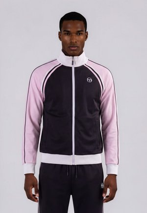 Homme portant une veste de sport zippée noire et rose avec col et poignets blancs, debout face à un fond neutre.