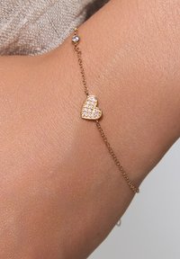 Złota bransoletka z zawieszką w kształcie serca. Serce zdobione jest małymi, przezroczystymi kamieniami szlachetnymi i jest przymocowane do delikatnego łańcuszka z małym akcentem w postaci koralików.