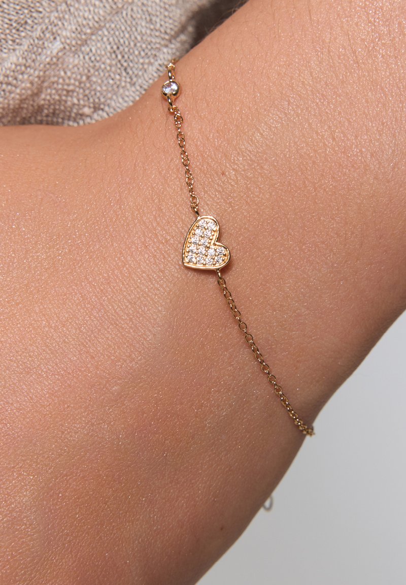 Złota bransoletka z zawieszką w kształcie serca. Serce zdobione jest małymi, przezroczystymi kamieniami szlachetnymi i jest przymocowane do delikatnego łańcuszka z małym akcentem w postaci koralików.