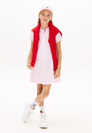 Ragazza che indossa un vestito polo rosa chiaro e un cappellino, con un maglione rosso appoggiato sulle spalle, calze bianche e scarpe da ginnastica bianche, in piedi con le gambe incrociate.