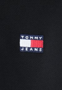 Tissu noir avec un petit patch rectangulaire du logo Tommy Jeans aux couleurs blanc, rouge et bleu marine et texte brodé.