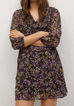 Frau mit langen braunen Haaren, die ein durchsichtiges schwarzes Kleid mit pinken, gelben und weißen Blumenmustern trägt, mit verschränkten Armen an einer schlichten Wand stehend.