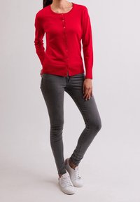 Červený cardigan s kulatým výstřihem a zapínáním na knoflíky vpředu, doplněný šedými přiléhavými džínami a bílými teniskami. Hladká textura.