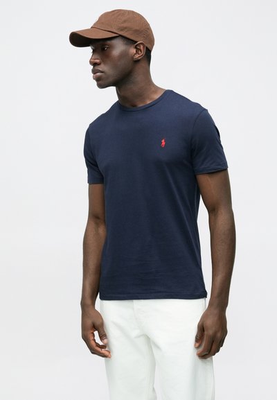 Tmavomodré tričko s krátkym rukávom z bavlny, s malým červeným logom na hrudi. Nosené s bledo-bielymi nohavicami a hnedou čiapkou.