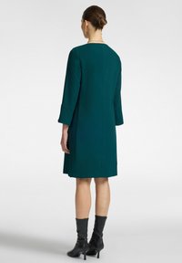 Tee-farbenes Kleid mit Dreiviertelärmeln und geradem Schnitt. Verfügt über einen runden Ausschnitt und einen glatten, strukturierten Stoff. Getragen mit knielangen schwarzen Stiefeln.