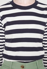 Camicia a maniche lunghe a righe con strisce orizzontali blu navy e bianche, scollo a girocollo e tessuto morbido. Abbinata a pantaloni verdi e un bottone marrone.