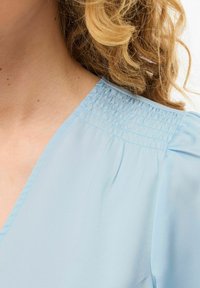 Blouse bleu clair avec un col en V et des détails froncés aux épaules. Le tissu est lisse avec un léger éclat.