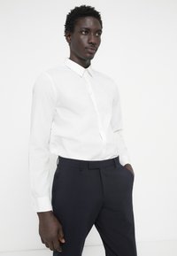 Bela srajca z dolgimi rokavi in gumbom ter s koničastim ovratnikom, v kombinaciji s temno modrimi hlačami. Gladek material z prilagojenim krojem, minimalistična zasnova.