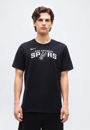 Jeune homme aux cheveux bruns courts portant un T-shirt noir des San Antonio Spurs, debout devant un fond clair uni.